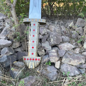 May include: A close-up of a sword with a gold-colored hilt and red gems. The hilt is decorated with intricate patterns. The sword is held against a background of rocks and greenery.
