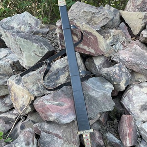 May include: A black leather scabbard with a gold-colored hilt and red gemstones. The scabbard is resting on a pile of rocks.