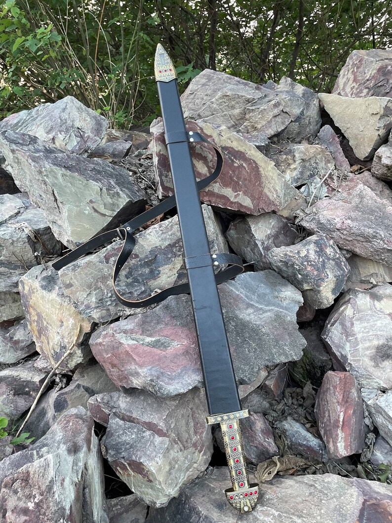 May include: A black leather scabbard with a gold-colored hilt and red gemstones. The scabbard is resting on a pile of rocks.