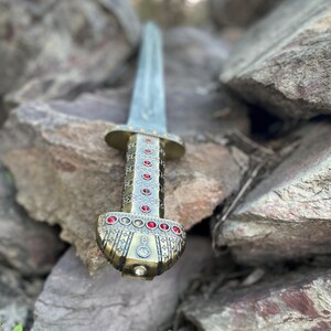 May include: A gold and silver sword with a red gem-encrusted hilt, resting on a pile of rocks.