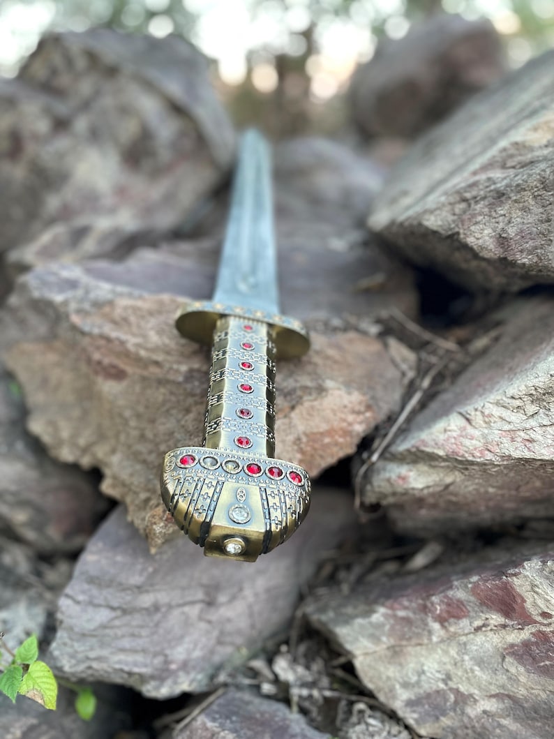 May include: A gold and silver sword with a red gem-encrusted hilt, resting on a pile of rocks.