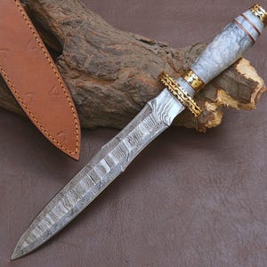 Könnte beinhalten: Ein feststehendes Messer mit einer gemusterten Damaststahlklinge, einem marmorierten weiß-grauen Griff und goldfarbenen Metallakzenten. Das Messer verfügt über ein verziertes Parierstück und eine passende hellbraune Lederscheide mit Nähten.