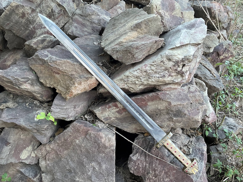 May include: A silver sword with a gold hilt and red gems. The sword is lying on a pile of rocks.