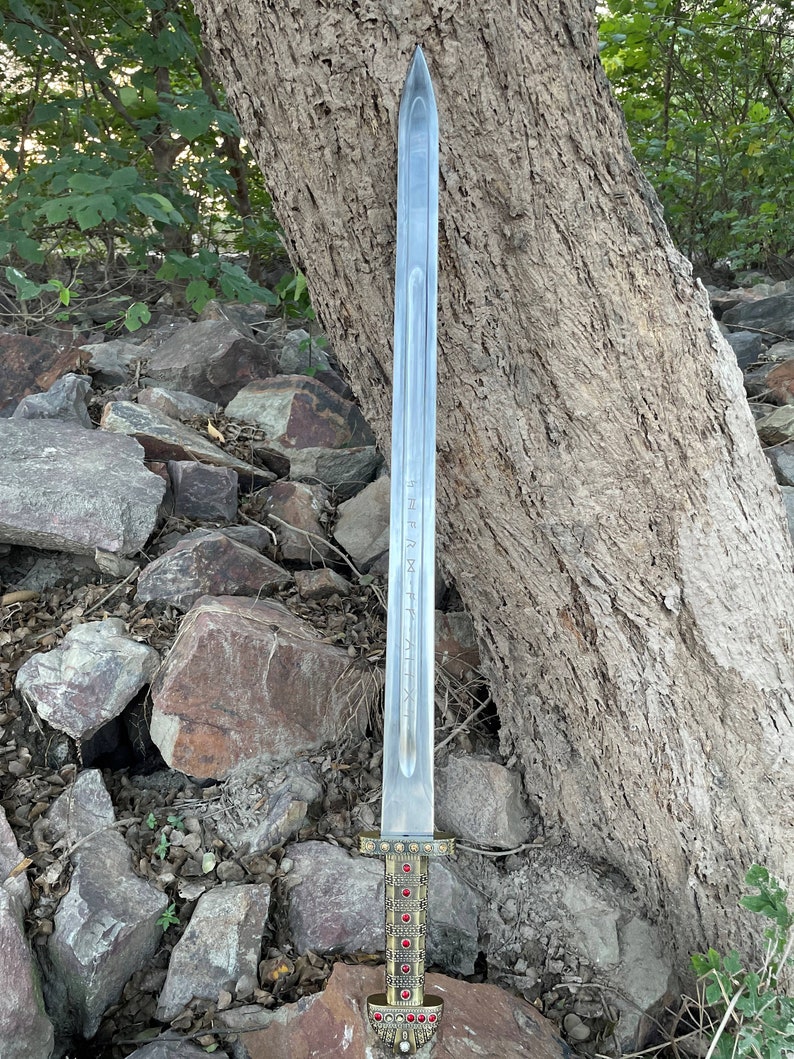 May include: A silver sword with a gold hilt and red gems. The sword is resting on a pile of rocks.