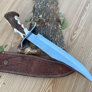 Puede incluir: Un cuchillo de caza con mango de asta de ciervo marrón y blanco y hoja plateada. El cuchillo está apoyado en un trozo de madera y una funda de cuero marrón.