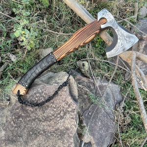 Könnte beinhalten: Eine handgefertigte Axt mit Holzgriff und Stahlklinge. Die Klinge hat ein eingraviertes Design. Der Griff ist mit schwarzem Leder umwickelt. Eine schwarze Kette ist am Griff befestigt. Die Axt liegt auf einem Felsen.