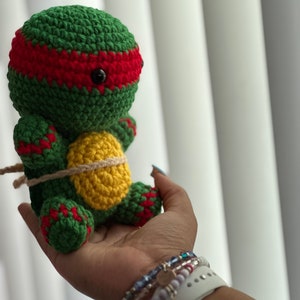 Könnte beinhalten: Eine handgefertigte Häkelpuppe einer Teenage Mutant Ninja Turtle, mit grünem Körper, roter Maske und gelbem Panzer. Die Puppe ist mit Bindfaden gebunden und hat schwarze Knopfaugen. Das Design ist ein süßes, weiches Spielzeug.