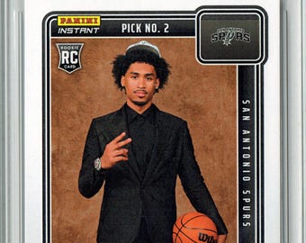 May include: A Rookie Card featuring Dylan Harper, a basketball player, holding a basketball. The card includes the text "DRAFT 2025" and "SAN ANTONIO SPURS". The card is encased in a protective holder.