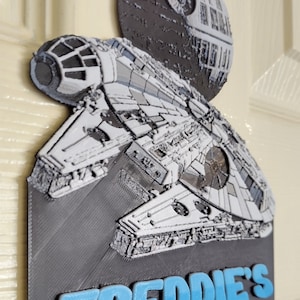 May include: A 3D printed door sign featuring a grey Millennium Falcon spaceship and a Death Star. The sign is on a white door and has the text "FREDDIE'S ROOM" in blue.