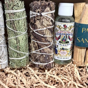 May include: A wooden box filled with sage bundles, a bottle of Florida Water cologne, and Palo Santo sticks. The sage bundles are wrapped with twine. The cologne bottle has a white cap and a colorful label. The Palo Santo sticks are bundled together.