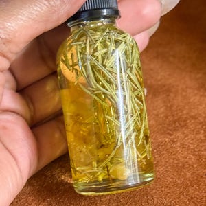 Puede incluir: Una botella de vidrio transparente llena de un l&iacute;quido amarillo y hierbas verdes. La botella tiene una tapa negra y se sostiene en una mano.