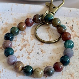 May include: A circular keychain featuring a series of smooth, multi-colored stone beads in shades of deep green, earthy brown, muted purple, and tan. The beads are strung on an elastic cord and attached to a bronze-toned metal lobster clasp and ring.