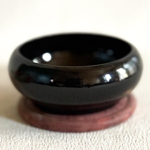 Puede incluir: Un cuenco negro brillante de forma redondeada, apoyado sobre una base circular de color marrón rojizo. La superficie del cuenco refleja la luz, creando reflejos. El diseño es simple y elegante.