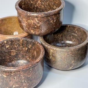 May include: Four stone bowls with a speckled, mottled pattern in shades of brown, tan, and beige. The bowls have a rounded shape with a slightly flared rim. The bowls are stacked and arranged in a close-up shot.