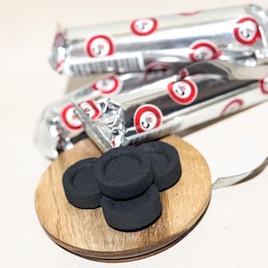 Op de afbeelding: Zwarte houtskoolbriketten op een klein houten bord. Op de achtergrond zilver verpakte houtskoolstokken met rood-witte cirkelvormige logo's. Een metalen gereedschap is ook zichtbaar.