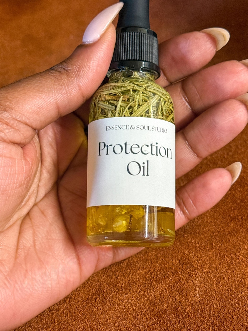 Puede incluir: Una botella de vidrio transparente con un gotero negro lleno de un l&iacute;quido amarillo y hierbas verdes. La botella tiene una etiqueta blanca que dice "Essence & Soul Studio Protection Oil".