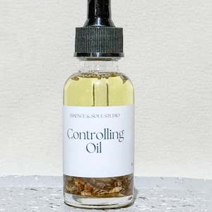 May include: A clear glass bottle of Controlling Oil with a black dropper top. The label reads "ESSENCE & SOUL STUDIO" and "Controlling Oil" in a minimalist font. The oil is light yellow, and the bottom of the bottle contains brown botanicals.