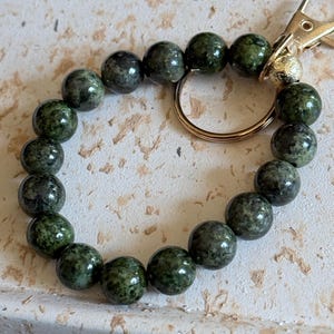 May include: A circular keychain featuring smooth, round, dark green mottled beads strung together. The keychain includes a gold-toned metal lobster clasp and a matching gold-toned circular ring attached to the bead strand for securing keys.