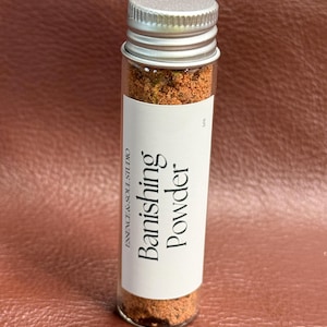 以下が含まれることがあります： 銀色の蓋が付いた小さなガラス瓶に、赤褐色の粉末が入っています。瓶のラベルには「Banishing Powder」と「Essence & Soul Studio」と書かれています。