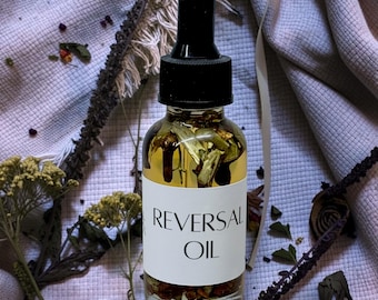 May include: A clear glass dropper bottle filled with a golden liquid and botanical pieces. The white label on the front features the black text REVERSAL OIL. The bottle has a black dropper cap and is positioned upright with visible plant matter inside.