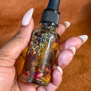 Puede incluir: Una botella de vidrio transparente con un gotero negro lleno de un aceite dorado y flores secas. Las flores son rojas, moradas y amarillas.