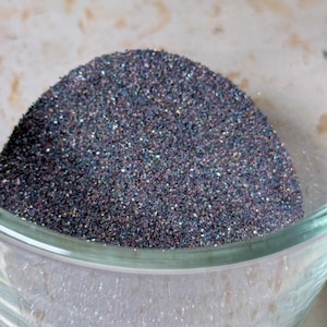 Puede incluir: Primer plano de un cuenco de vidrio transparente lleno de un montón de purpurina oscura e iridiscente. Las partículas de purpurina son una mezcla de púrpura oscuro, azul y plata, creando un efecto brillante. El cuenco está sobre una superficie clara.