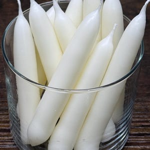 May include: A collection of white taper candles with pointed tips, standing upright inside a clear glass cylinder. The candles are smooth, cylindrical, and arranged in a cluster, filling the glass container with their uniform white wax appearance.