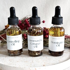 Puede incluir: Tres frascos cuentagotas de vidrio con tapas negras y etiquetas blancas. Las etiquetas dicen "Controlling Oil", "Command & Conquer Oil" y "Domination Oil". Los frascos están llenos de aceite y rodeados de flores y hierbas secas.