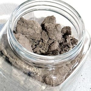 May include: A clear glass jar filled with dark brown, irregularly shaped pieces. The jar has a wide mouth and a square base. The contents appear to be a dried, earthy material. The jar is sitting on a light-colored surface.