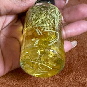 Puede incluir: Una botella de vidrio transparente llena de un l&iacute;quido amarillo y hierbas verdes. La botella tiene una tapa negra.