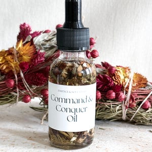 May include: A clear glass bottle with a black dropper top, filled with oil and botanicals. A white label reads "Command & Conquer Oil." Dried flowers and herbs in shades of red, orange, and green are in the background.