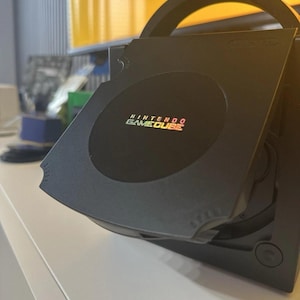 Puede incluir: Una consola de videojuegos Nintendo GameCube negra con una unidad de disco de carga superior y la tapa abierta. El centro circular de la tapa muestra el logotipo de Nintendo GameCube en texto blanco y naranja. Incluye un asa de transporte.