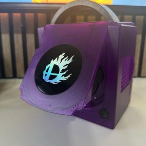 Könnte beinhalten: Eine durchscheinende violette Videospielkonsole in quadratischer Form mit einem Griff an der Oberseite. Die Disc-Abdeckung ist geöffnet und gibt den Blick auf ein rundes schwarzes Emblem mit einem weißen, von Flammen umgebenen Smash Bros-Logo frei.