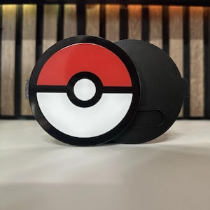 Puede incluir: Un emblema circular de Poké Ball en negro, rojo y blanco con una banda central negra y un círculo blanco. El emblema está unido a una base circular negra sólida con un acabado mate suave, presentando un diseño limpio y funcional.