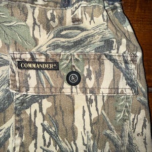 K&ouml;nnte beinhalten: Nahaufnahme einer Camouflage-Hose mit einer Tasche mit dem Wort "COMMANDER". Die Hose hat einen Knopfverschluss und einen Kordelzug in der Taille. Das Tarnmuster umfasst Braunt&ouml;ne, Gr&uuml;n und Grau.