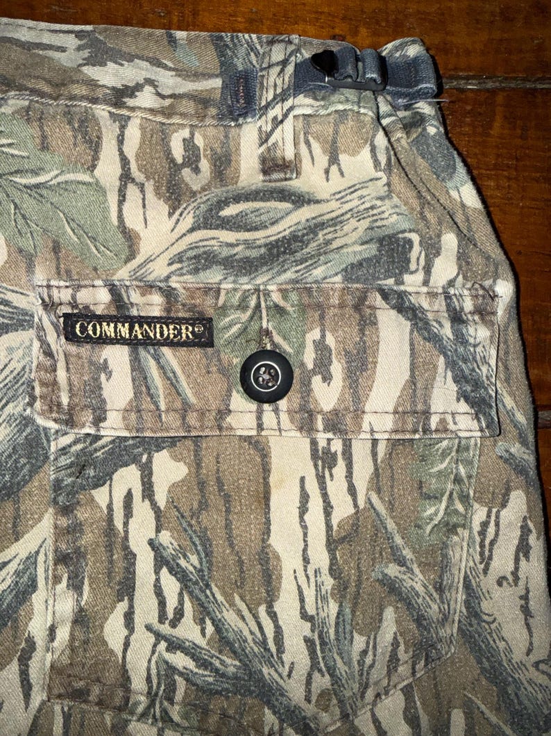 K&ouml;nnte beinhalten: Nahaufnahme einer Camouflage-Hose mit einer Tasche mit dem Wort "COMMANDER". Die Hose hat einen Knopfverschluss und einen Kordelzug in der Taille. Das Tarnmuster umfasst Braunt&ouml;ne, Gr&uuml;n und Grau.