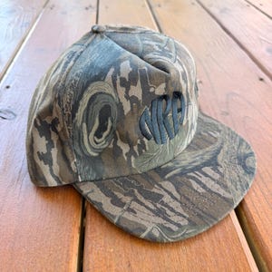 May include: Camouflage print baseball cap with the embroidered letters "NRPA" in dark green. The cap features a flat brim and a structured crown. The hat is made of a durable fabric and is designed for outdoor wear.