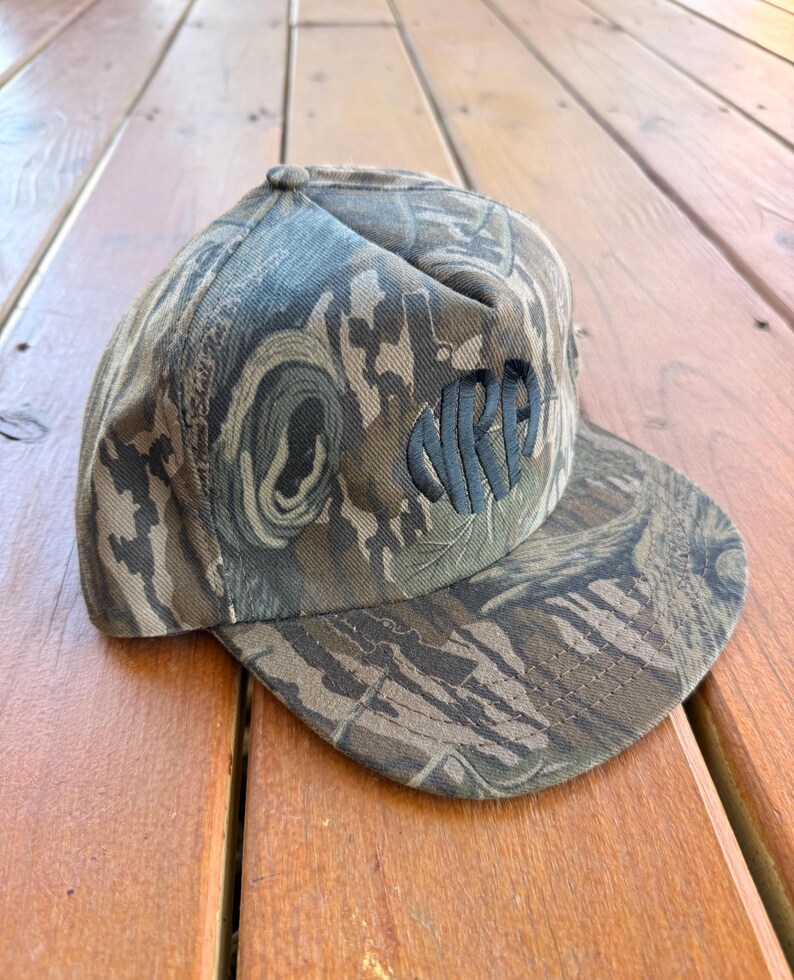 May include: Camouflage print baseball cap with the embroidered letters "NRPA" in dark green. The cap features a flat brim and a structured crown. The hat is made of a durable fabric and is designed for outdoor wear.