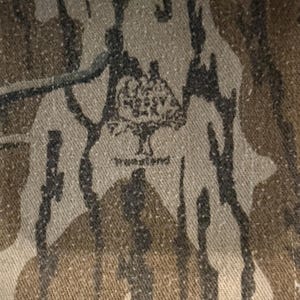 K&ouml;nnte beinhalten: Nahaufnahme eines Camouflage-Stoffs mit einem Muster aus braunen, schwarzen und beigen Formen. Der Stoff hat das Mossy Oak True Land Logo aufgedruckt. Das Design erinnert an einen Wald.