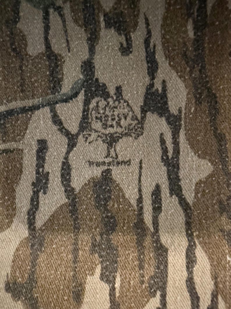 K&ouml;nnte beinhalten: Nahaufnahme eines Camouflage-Stoffs mit einem Muster aus braunen, schwarzen und beigen Formen. Der Stoff hat das Mossy Oak True Land Logo aufgedruckt. Das Design erinnert an einen Wald.
