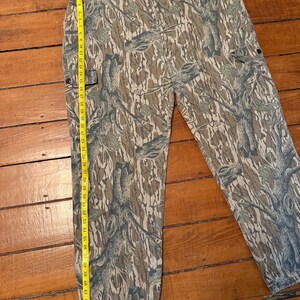 K&ouml;nnte beinhalten: Tarnfarbene Cargohose in Braun-, Gr&uuml;n- und Graut&ouml;nen. Die Hose hat mehrere Taschen und einen Knopfverschluss. Ein Ma&szlig;band zeigt eine L&auml;nge von etwa 119 cm an.
