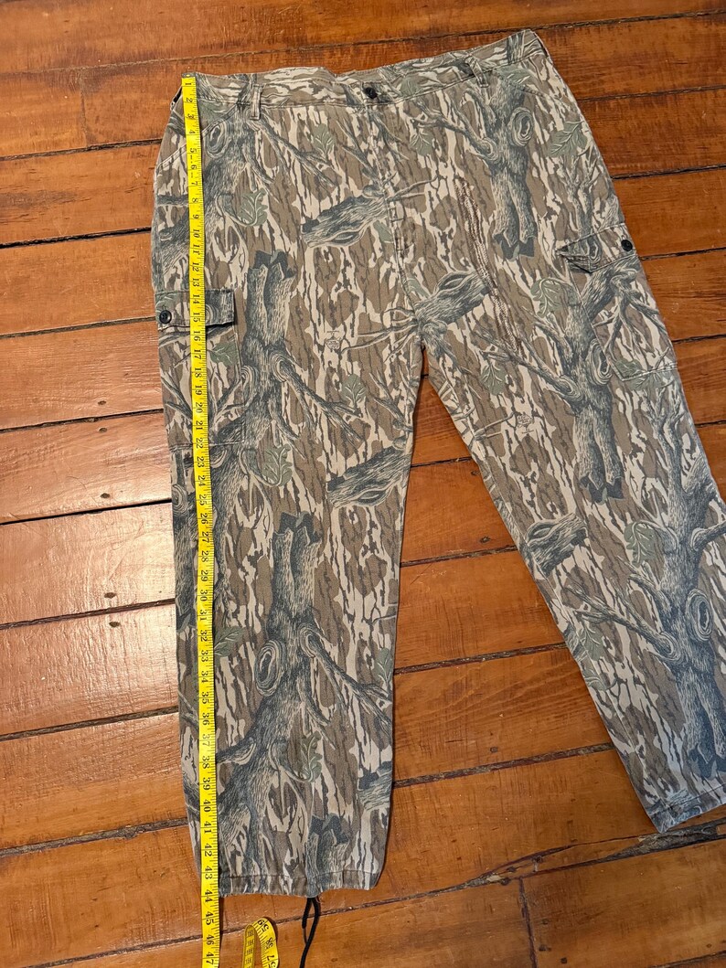 K&ouml;nnte beinhalten: Tarnfarbene Cargohose in Braun-, Gr&uuml;n- und Graut&ouml;nen. Die Hose hat mehrere Taschen und einen Knopfverschluss. Ein Ma&szlig;band zeigt eine L&auml;nge von etwa 119 cm an.