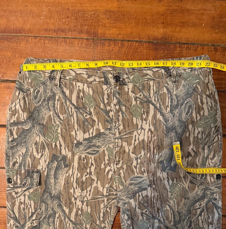 K&ouml;nnte beinhalten: Tarnfarbene Cargohose mit Knopfverschluss und zwei Vordertaschen. Ein gelbes Ma&szlig;band zeigt eine Taillenmessung von ca. 58 cm. Die Hose hat ein braunes, gr&uuml;nes und beiges Baumrindenmuster.