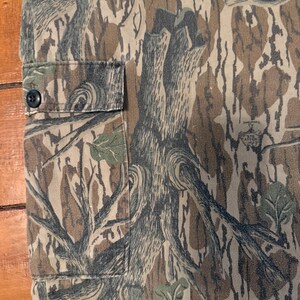 K&ouml;nnte beinhalten: Nahaufnahme eines Camouflage-Stoffs mit einem Muster aus braunen, gr&uuml;nen und grauen baumartigen Formen. Auf der linken Seite des Stoffes ist eine kleine Tasche mit einem schwarzen Knopf zu sehen. Der Stoff scheint Teil eines Kleidungsst&uuml;cks zu sein.