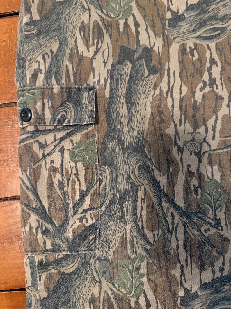 K&ouml;nnte beinhalten: Nahaufnahme eines Camouflage-Stoffs mit einem Muster aus braunen, gr&uuml;nen und grauen baumartigen Formen. Auf der linken Seite des Stoffes ist eine kleine Tasche mit einem schwarzen Knopf zu sehen. Der Stoff scheint Teil eines Kleidungsst&uuml;cks zu sein.