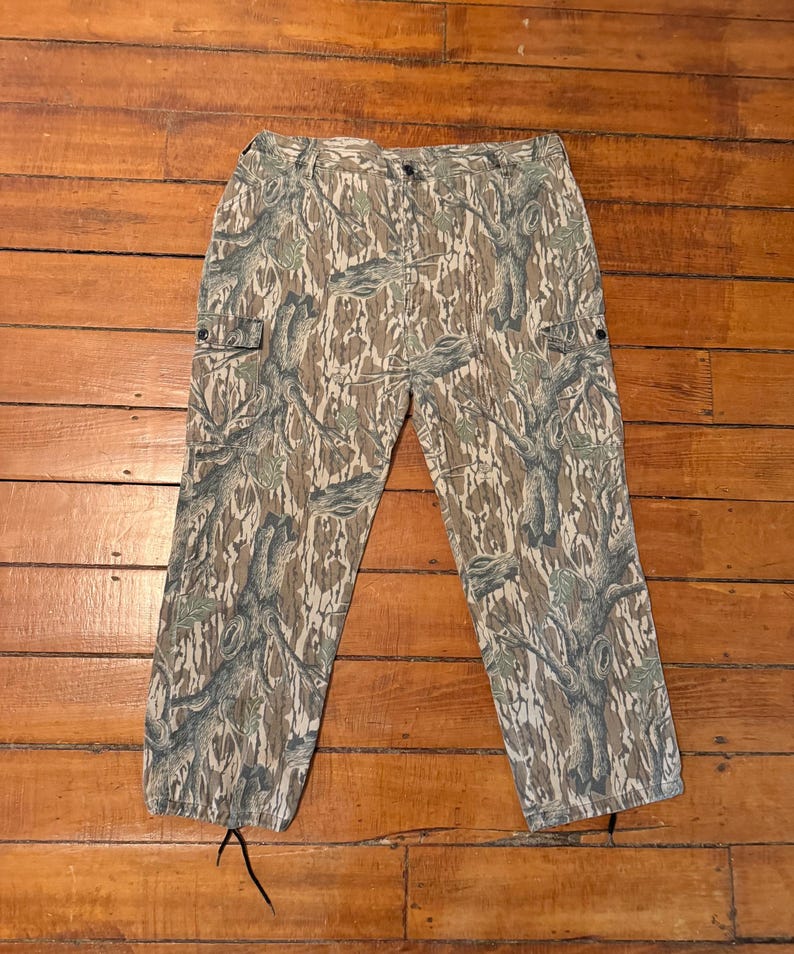 K&ouml;nnte beinhalten: Tarnfarbene Cargohose in Braun-, Gr&uuml;n- und Graut&ouml;nen. Die Hose hat mehrere Taschen und einen Kordelzug am unteren Saum. Der Stoff hat ein Baumrindenmuster.