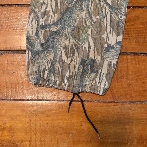 K&ouml;nnte beinhalten: Nahaufnahme einer Camouflage-Hose mit Kordelzug. Die Hose hat ein braunes, gr&uuml;nes und graues Baumrindenmuster. Der Kordelzug ist schwarz. Die Hose liegt auf einer Holzoberfl&auml;che.