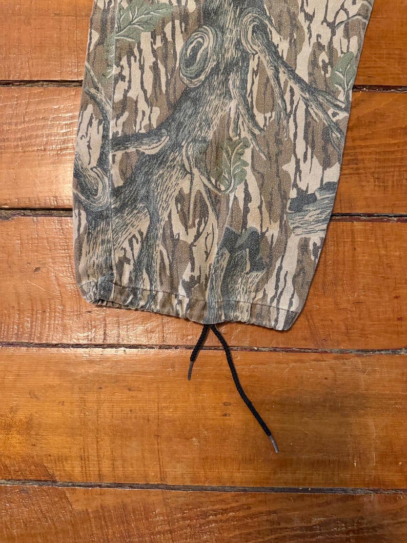 K&ouml;nnte beinhalten: Nahaufnahme einer Camouflage-Hose mit Kordelzug. Die Hose hat ein braunes, gr&uuml;nes und graues Baumrindenmuster. Der Kordelzug ist schwarz. Die Hose liegt auf einer Holzoberfl&auml;che.