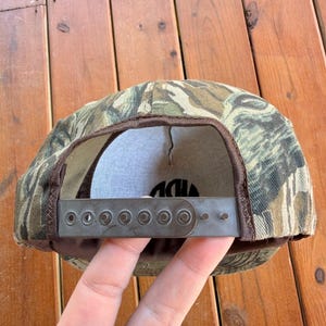May include: Camouflage baseball cap with a brown brim and adjustable strap. The cap features a green, brown, and tan pattern. The interior is white with black text. The adjustable strap is gray metal.