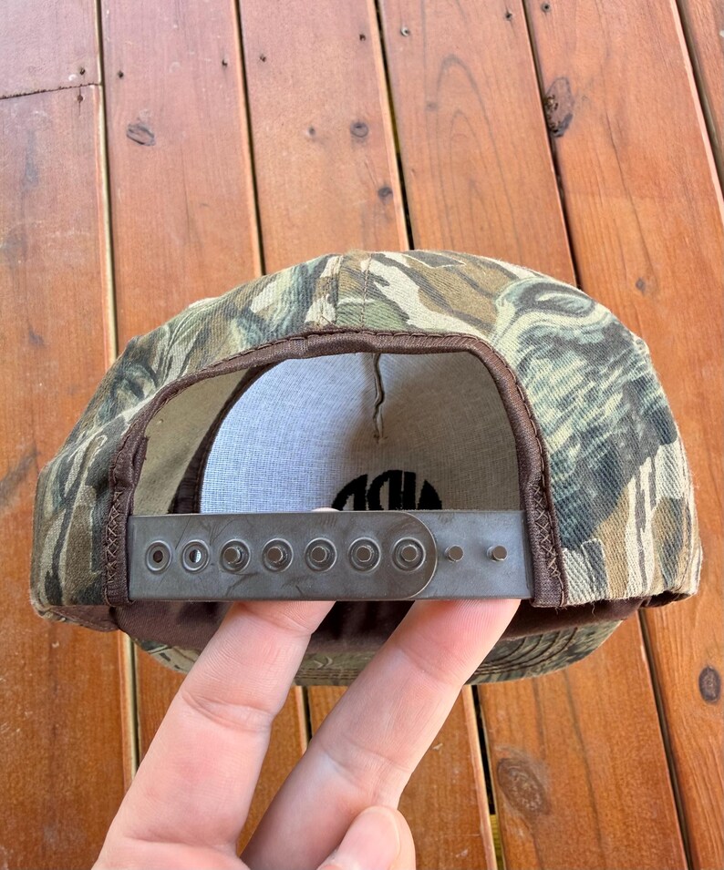 May include: Camouflage baseball cap with a brown brim and adjustable strap. The cap features a green, brown, and tan pattern. The interior is white with black text. The adjustable strap is gray metal.